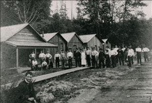 A large group of Japanese men stand outside wooden buildings with forest in the background